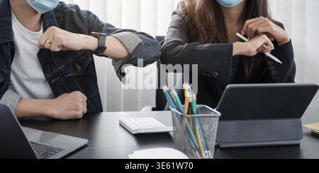 Zwei junge, vielfältige Geschäftspartner tragen Gesichtsschutzmasken, stoßen Ellbogen an und grüßen einander während der Arbeit während covid 19 Quarantäne Stockfoto
