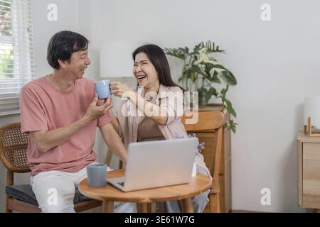 Ein glückliches älteres Paar teilt sich einen freudigen Toast mit ihren Kaffeetassen, während es in seinem komfortablen Zuhause Online-Aktivitäten genießt Stockfoto