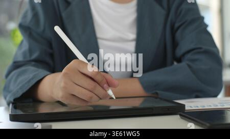Eine Frau schreibt mit einem weißen Stift auf eine Tablette. Sie trägt eine blaue Jacke und ein weißes Hemd Stockfoto