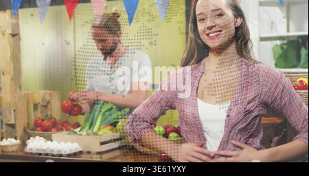 Lächelnde Frau in Karo, die vor dem Stand steht, Kollege arrangiert Tomaten in Holzkisten Stockfoto
