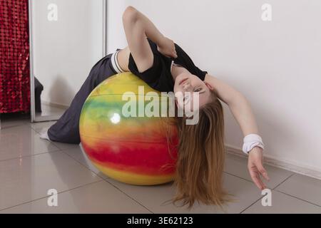 Eine junge Frau lehnt sich auf einen lebendigen Trainingsball zurück, während sie in einem gut beleuchteten Studio eine Stretchroutine durchführt. Sie konzentriert sich auf Balance und Flexibilität Stockfoto
