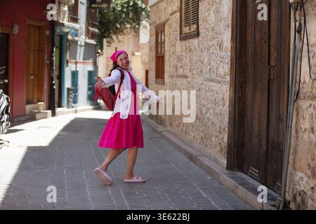 Eine Erwachsene Frau in rosa Kleid und passendem Hut spaziert spielerisch über eine enge Kopfsteinpflasterstraße und genießt die Umgebung in einer malerischen Historica Stockfoto