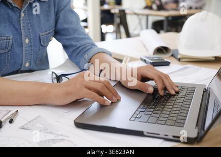 Nahaufnahme eines Architekten und Ingenieur, der einen Laptop benutzt, während er die Blaupausen überprüft. Ein professioneller Arbeitsbereich für die Projektplanung Stockfoto