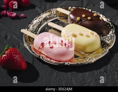 Essen, drei handwerklich hergestellte Eissorten sitzen auf einem Silberteller, mit leuchtenden Farben und Belägen. Frische Erdbeeren und Himbeeren werden um t gelegt Stockfoto