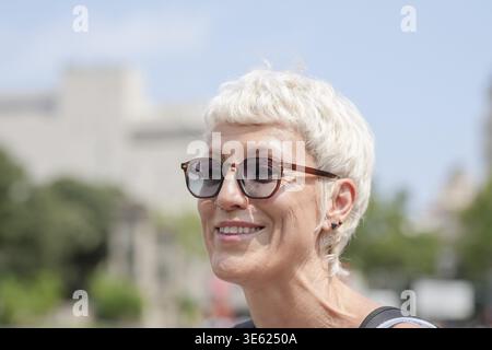 Eine Frau mit kurzen Haaren und Brille lächelt. Sie trägt ein schwarzes Kleid und eine Sonnenbrille. Die Szene spielt in einer Stadt mit einem Gebäude in der BA Stockfoto