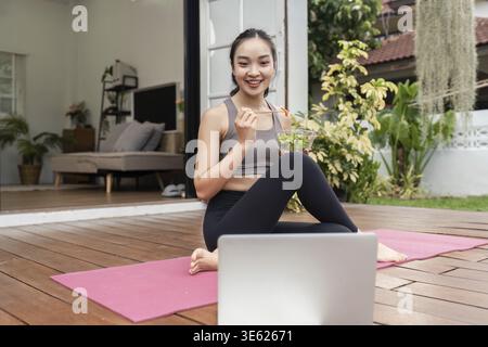 Eine junge Frau sitzt auf einer Yogamatte, isst einen Salat und lächelt ihren Laptop an, fördert gesunde Ernährung und Wohlbefinden Stockfoto
