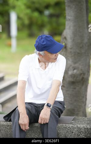 Ein älterer Mann mit blauem Hut und weißem Hemd sitzt auf einer Bank. Er trägt eine Uhr und hat einen grauen Bart Stockfoto