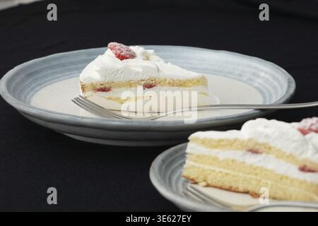 Ein kleiner japanischer Erdbeer-Shortcake und je eine Gabel auf zwei stilvollen, großen und kleinen Tellern im japanischen Stil auf einem schwarzen Tuch auf einem weißen Tisch Stockfoto