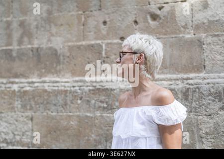 Eine Frau mit kurzen Haaren und Brille steht vor einer Steinmauer. Sie lächelt und blickt in den Himmel Stockfoto