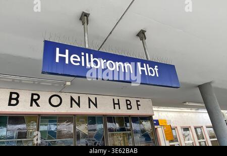 Wirtschaft in Heilbronn, Baden-Württemberg 28.03.2026, Wirtschaft v.l. Symbolbild, Beispielbild, Haltepunkt, Zughalt, Standort, Bahnhofsschild, Ortskennzeichnung Hinweisschild Außenansicht Hauptbahnhof Heilbronn dieses Bild zeigt ein Detail einer Bahnsteigausstattung am Heilbronner Hauptbahnhof. Zentrum des Bilds: Ein blaues Bahnhofsschild mit der weißen Aufschrift Heilbronn Hbf in der serifenlosen Standardschrift der Deutschen Bahn.Halterung: Das Schild ist an einem grauen Metallmast befestigt, der vertikal durch die Bildmitte verläuft.Sicherheitstechnik: Direkt unter dem Schild ist eine wei Stockfoto