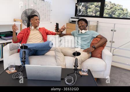 Aufnahme verschiedener männlicher Freunde, die auf dem Sofa im Podcast-Studio plaudern, mit Laptop und Mikrofonen Stockfoto
