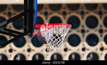Basketball, der durch einen roten Korb und ein Netz auf einem Platz im Freien mit verschwommenem Hintergrund schwingt Stockfoto