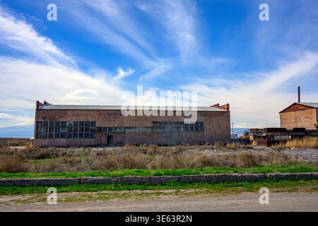 Ein großes Industriegebäude aus Ziegelstein steht unter hellblauem Himmel stehen gelassen. Zerbrochene Fenster und bewachsenes Gras umgeben die verfallene Struktur. Beton p Stockfoto