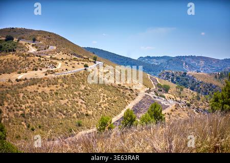 Eine malerische Aussicht auf sanfte Hügel, bedeckt mit trockenem goldenem Gras und karger Vegetation unter einem klaren blauen Himmel. Eine kurvige asphaltierte Straße schlängelt sich entlang Stockfoto