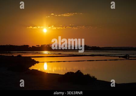 Goldener Sonnenuntergang über Salzwasserbecken an der Küste mit spiegelähnlichen Sonnenspiegeln auf ruhigen Wasseroberflächen. Die helle Sonne sitzt tief in einem orange-bernsteinfarbenen Stockfoto
