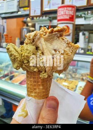 Ein Nahfoto einer Hand, die einen Waffelkegel mit zwei großzügigen Schaufeln handgemachtem Eis hält. Ein Messlöffel ist pistazienaromatisiert mit einem unverwechselbaren Stockfoto