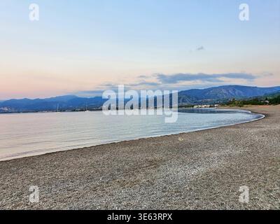 Eine ruhige Küstenlandschaft in der Dämmerung mit einem gekrümmten Kiesstrand im Vordergrund mit sanften Wellen am Ufer. Das ruhige Meer Stockfoto