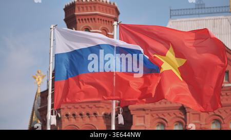 Fahnen von Russland und Vietnam winken zusammen vor einem historischen roten Backsteingebäude vor einem blauen Himmel. Stockfoto