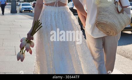 Romantisches Paar, das durch die Sommerstraße spaziert, Lotusblumen hält und Hochzeitskleidung trägt Stockfoto