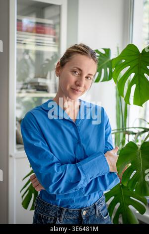 Ruhige glückliche Frau, die neben dem Garten steht, bei Monstera-Blättern, Pflanzenpflege Stockfoto