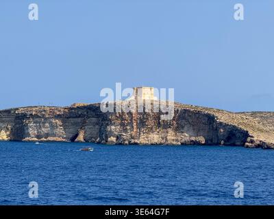 Blick auf den Turm von Santa Maria auf der Insel Comino über zerklüfteten Kalksteinklippen, gesehen über das tiefblaue Mittelmeer unter klarem Himmel. Stockfoto