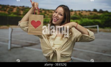 Frau zeigt mit dem Finger auf die Papiertüte mit rotem Herzen auf dem Tennisplatz, hält den Griff und lächelt breit; liebe Freude. Stockfoto