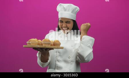 Die junge Küchenchefin in weißer Uniform hält ein hölzernes Tablett mit Muffins und pumpt im Studio nach dem Backsieg die Faust. Stockfoto