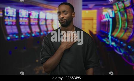 Junger afroamerikanischer Mann mit Hand auf der Brust in der Nähe von beleuchteten Spielautomaten im Eingangsbereich des Casinogebäudes; bedauern Reue-Entschuldigung. Stockfoto