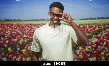 Der Mann lächelt und stellt die Brille mit der rechten Hand ein, trägt ein blasses T-Shirt, während er auf einem Feld zwischen Tulpenreihen steht, nach rechts gerichtet; Ruhe. Stockfoto