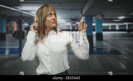 Mittelalter hispanische blonde Frau in weißem Hemd berührt ihr Ohr mit der linken Hand und zeigt den rechten Zeigefinger nach oben zwischen Betonsäulen und geparkten c Stockfoto