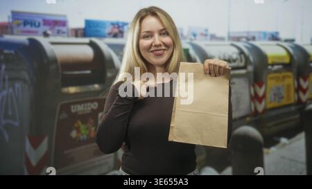 Eine blonde, lächelnde Frau, die eine braune Papiertüte hält, zeigt auf der städtischen Straße mit dem Finger auf die Brust, indem sie Abfalleimer recycelt; Selbstvertrauen stärkt. Stockfoto