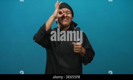 Küchenchef in schwarzer Uniform, der Metallspachtel hält und im teelfarbenen Studio ein gutes Zeichen über dem Auge macht; verspielte Freude. Stockfoto