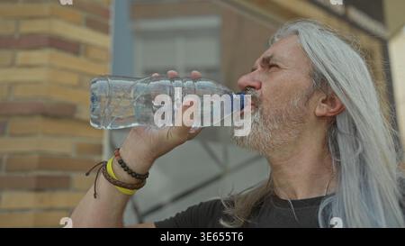 Mann, der aus einer klaren Plastikflasche trinkt, von Hand in Mund, in der Nähe eines Ziegelgebäudes auf der Straße steht; Erfrischungsgetränk. Stockfoto