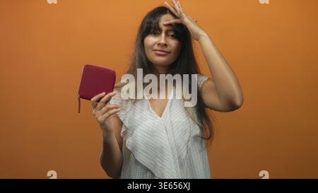 Junge Latina-Brünette, die mit der Hand bis zur Stirn eine rosa Brieftasche in einem orangen Studio hält; Müdigkeit. Stockfoto