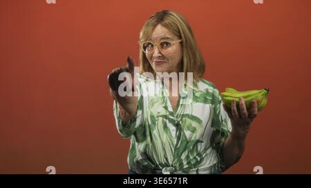 Mittelalter hispanische Frau mit blondem Haar und Brille zeigt den Finger zur Kamera, während sie im Studio Bananen hält; spielerisches Selbstvertrauen. Stockfoto