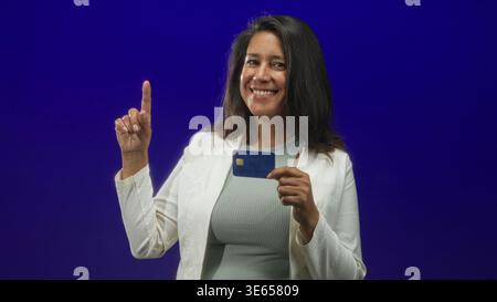 Frau, die Kreditkarte und Zeigefinger hält, während sie im Studio lächelt; Vertrauen in finanzielle Stärkung. Stockfoto