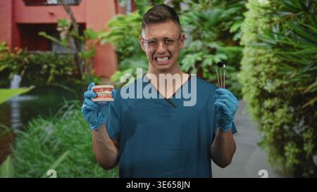 Männlicher Zahnarzt in blauen Peelings, Zahnmodell und Spiegel mit Handschuhen halten, Schielen im Bauwerk; professionelle Betreuung. Stockfoto