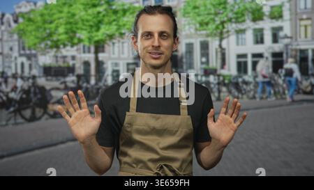 Junger Mann mit Händen, die Palmen auf der Straße hochgehoben haben, mit Schürze und schwarzem Hemd, langen Haaren, die zurückgebunden sind; Entschuldigung Demut. Stockfoto