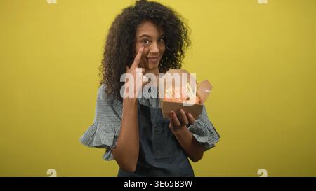 Teenager-Frau mit lockigem Haar in der Schürze zeigt Finger an Wange, während sie eine Kiste mit gebratenem Huhn und Pommes vor gelbem Studio-Hintergrund hält; S. Stockfoto