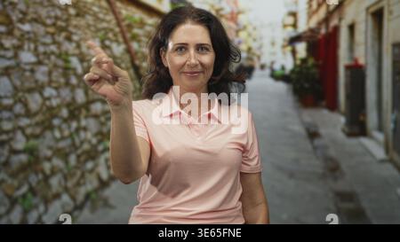 Frau zeigt mit dem Zeigefinger in einer engen Steinstraße mit subtilem Lächeln und direktem Blick; ruhiges Selbstvertrauen. Stockfoto