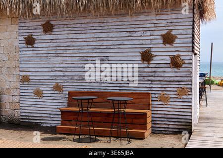 Sitzanordnung aus Holz mit zwei schwarzen Stühlen vor einer weißen Holzwand mit gewebten Schildkröten- und Krabbenmotiven in der Nähe eines Sandstrandes Stockfoto