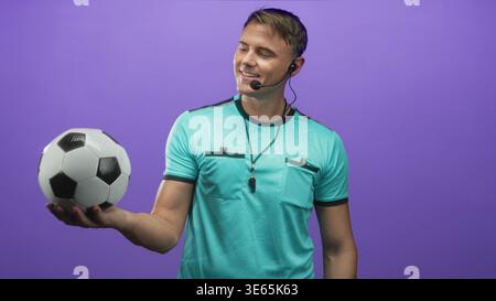 Der Schiedsrichter hält den Fußball in gestreckter Hand, mit Pfeife und Headset vor der violetten Studiowand; Selbstvertrauen. Stockfoto