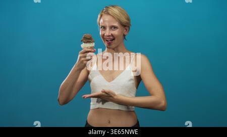 Weißblonde Frau hält Eisbecher nahe dem Gesicht, während sie mit geschlossenen Augen im Studio vor blauem Hintergrund lächelt; Freude am Sommervergnügen. Stockfoto