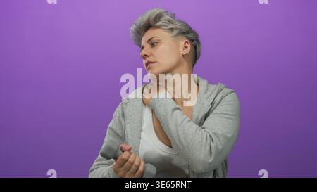 Frau mit kurzen Haaren in grauem Hoodie, die Hantel hält und den Hals im lila Studio umklammert; Unwohlsein. Stockfoto