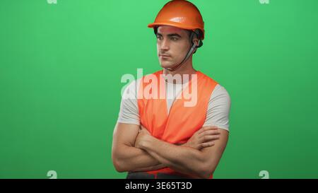 Junger Ingenieur mit orangefarbenem Schutzhelm und Sicherheitsweste mit gekreuzten Armen und Headset im grünen Studio; Vertrauensdienst. Stockfoto