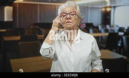 Eine ältere Frau mit weißem Hemd passt eine bunte Brille an und zeigt den Finger auf den Tempel in einem Gebäude; Entschlossenheit Weisheit. Stockfoto