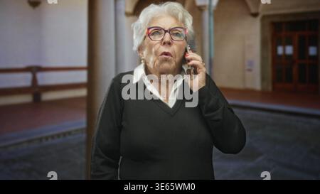 Seniorin hält Smartphone mit sichtbarer Hand und Brille im Foyer des Gebäudes; Sorge um das Altern. Stockfoto