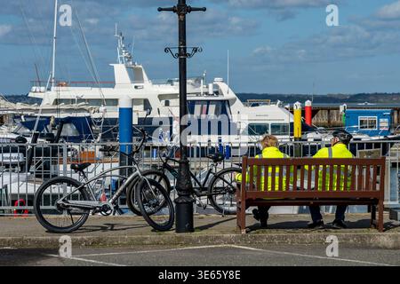Ältere Paare in gut sichtbarer Fahrradkleidung auf einer Bank sitzen und sich von ihrer Fahrradtour in Yarmouth auf der isle of wight, Großbritannien, erholen Stockfoto
