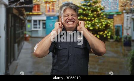 Polizist bedeckte Ohren mit Fingern, grimperte auf einer nassen Fußgängerzone neben einem beleuchteten weihnachtsbaum und Ladenfronten; Feiertagsstress. Stockfoto