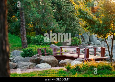 Eine friedliche japanische Gartenszene mit einer kleinen Holzbrücke in rötlich-brauner Farbe, umgeben von großen grauen Felsen, grünem Gras und Bäumen wi Stockfoto